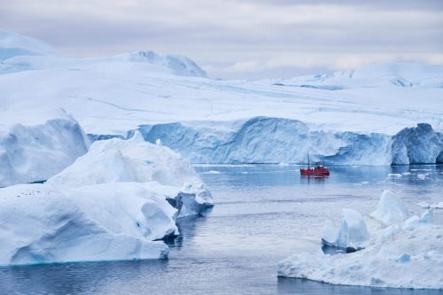 Floating icebergs in Ilulissat with a small red boat among pale blue ice under a cloudy sky.