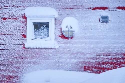 Close view of a frost-covered red wooden wall in Sisimiut with a small window and exterior fixtures encased in ice.
