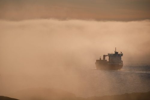 Ship partially obscured by dense fog in coastal waters near Sisimiut, with muted orange light diffused through the mist.