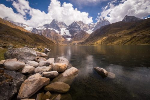Mountain lake in Peru with rocks in the foreground and steep peaks reflected in still water.