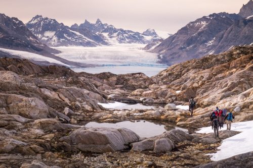 People walking through rocky terrain in Greenland with a glacier filling the background.