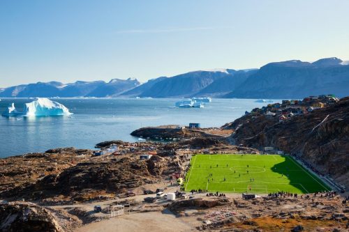 Rocky coastal landscape near Uummannaq with a bright green sports field beside the sea and icebergs in the distance.