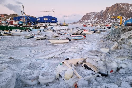 Icy shoreline in Sisimiut harbor with fishing boats, equipment, and snow-covered ground.