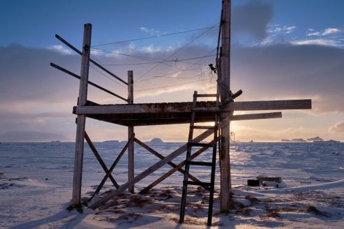 Simple wooden structure standing on frozen ground near Qaanaaq with sea ice and distant mountains
