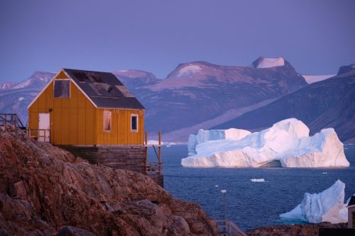 Yellow house on rocky ground overlooking the sea near Uummannaq, with icebergs floating in cold blue water.