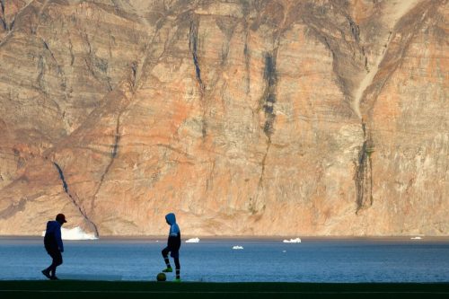 Two kids playing soccer in front of a steep rock face in Uummannaq.