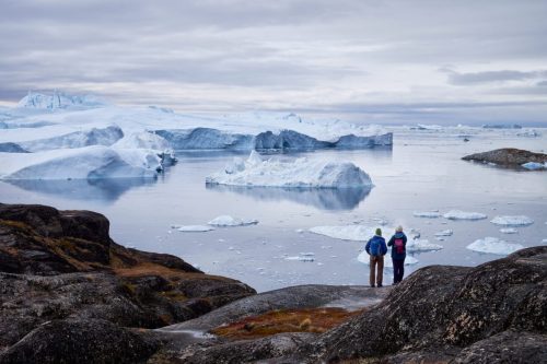 Two figures standing on rocky ground overlooking floating icebergs in Ilulissat Icefjord.